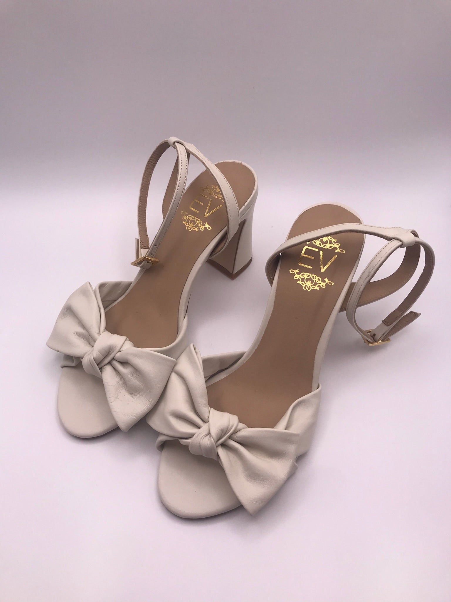 Sandálias de senhora em pele creme com laço frontal EV; sapatos de salto alto grosso com tira no tornozelo e fivela dourada; calçado elegante para noivas ou cerimónias especiais